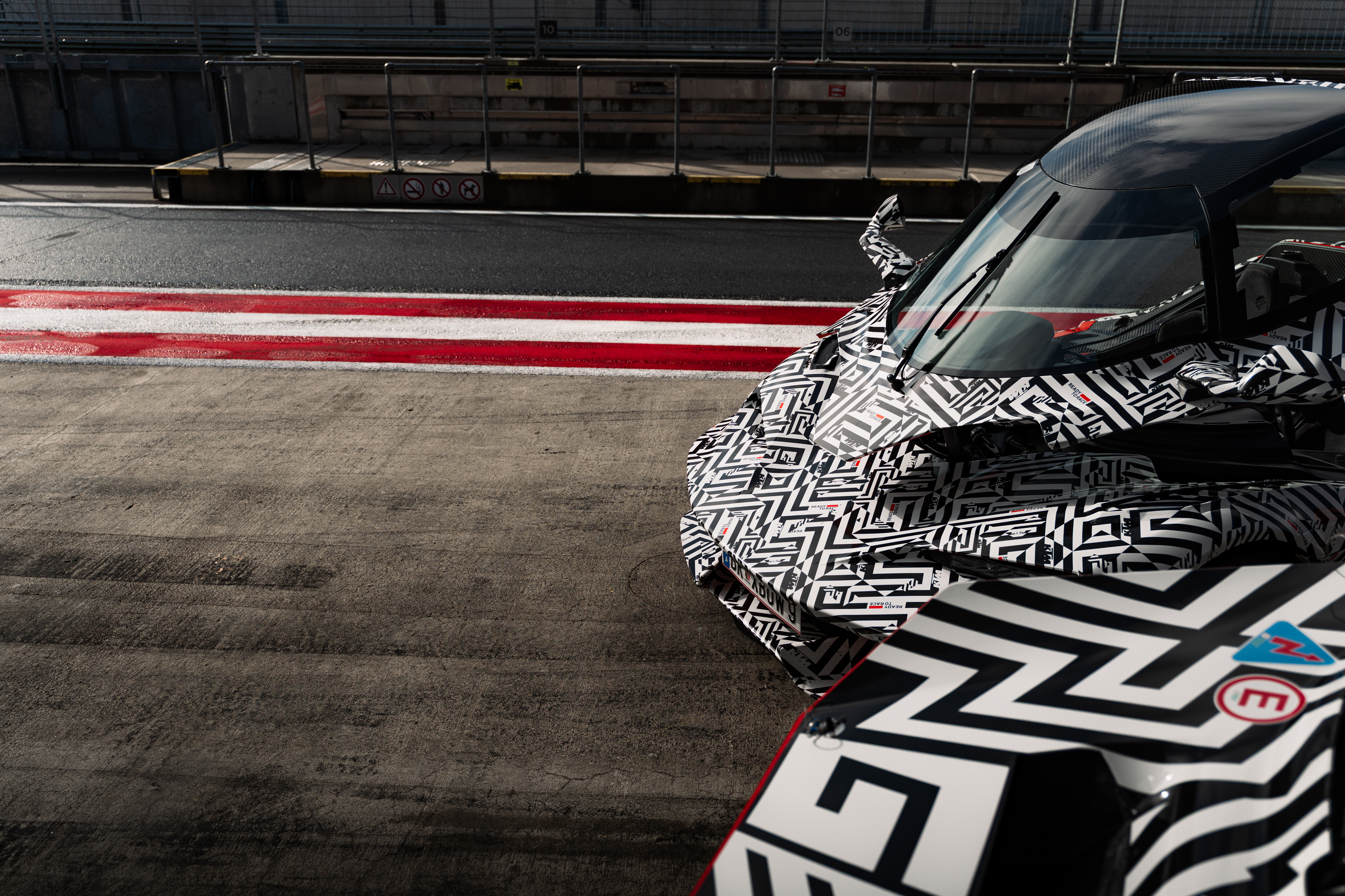 A sports car with a black and white geometric pattern parked on a race track beside red and white lines, under bright sunlight.