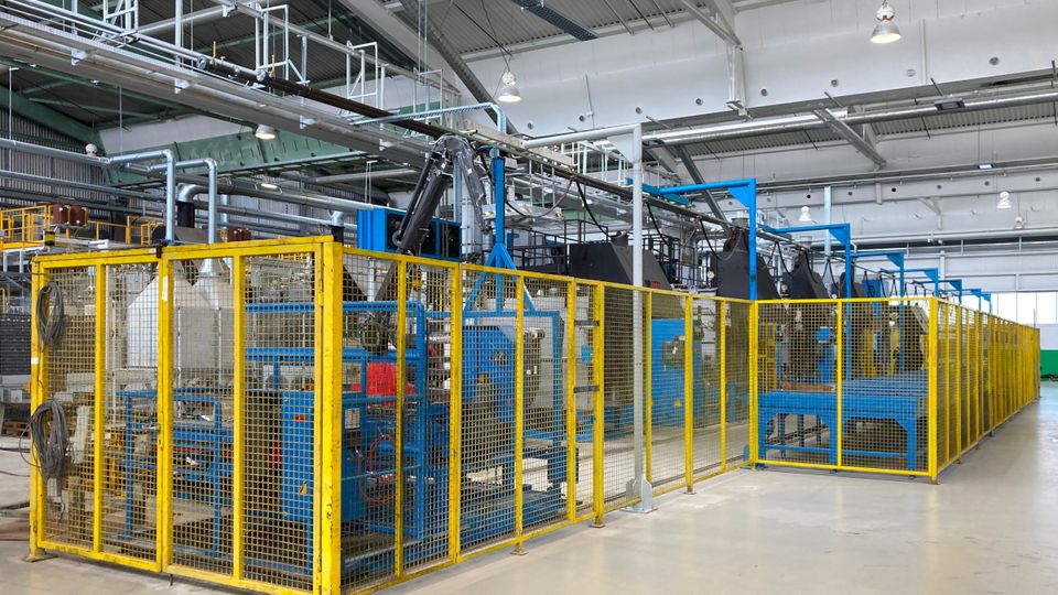 A large industrial factory interior with machinery enclosed by yellow safety barriers and overhead lights illuminating the space.