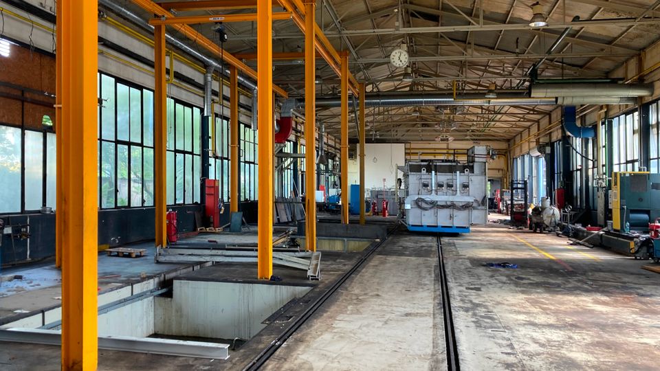 An empty industrial warehouse with yellow beams, large windows, and machinery in the background under a high ceiling.