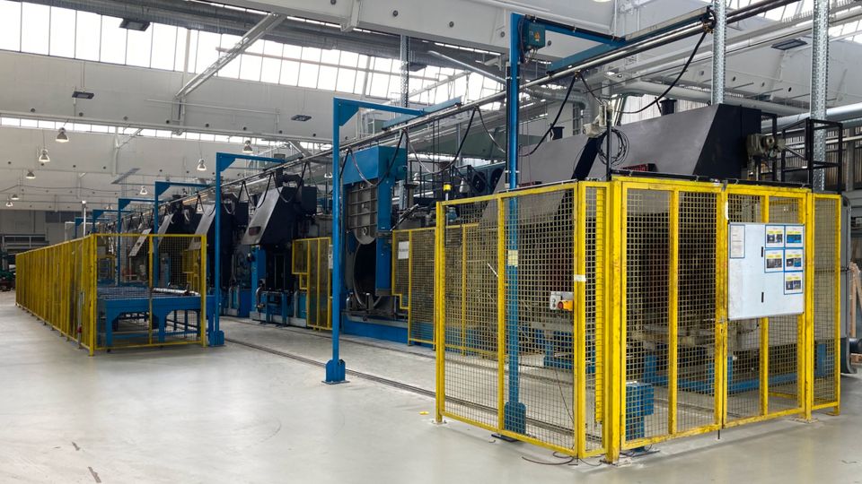 Modern industrial factory interior with metal machines enclosed by yellow safety fences, under a ceiling with large skylights.