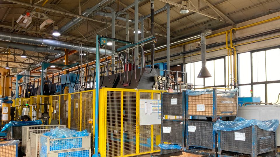 Interior of a factory with machinery, metal cages, and boxes covered in blue plastic, under a high ceiling with exposed pipes and ducts.