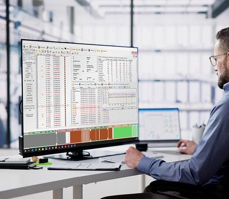 A man in an office setting works on a large computer monitor displaying complex data and graphs, with a tablet beside him.