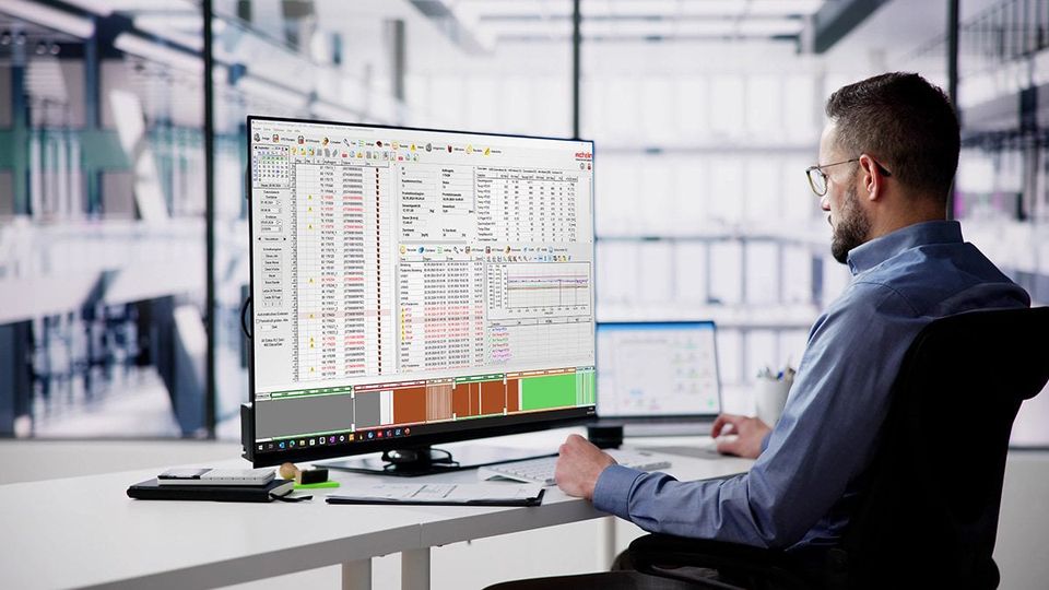 A man in an office setting works on a large computer monitor displaying complex data and graphs, with a tablet beside him.