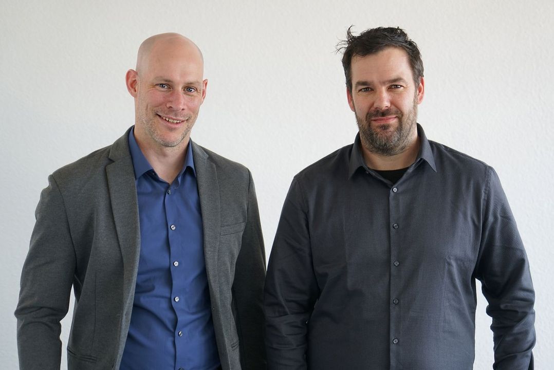 Two men standing side by side, both wearing button-up shirts; one in a gray blazer. They are smiling in front of a plain white background.