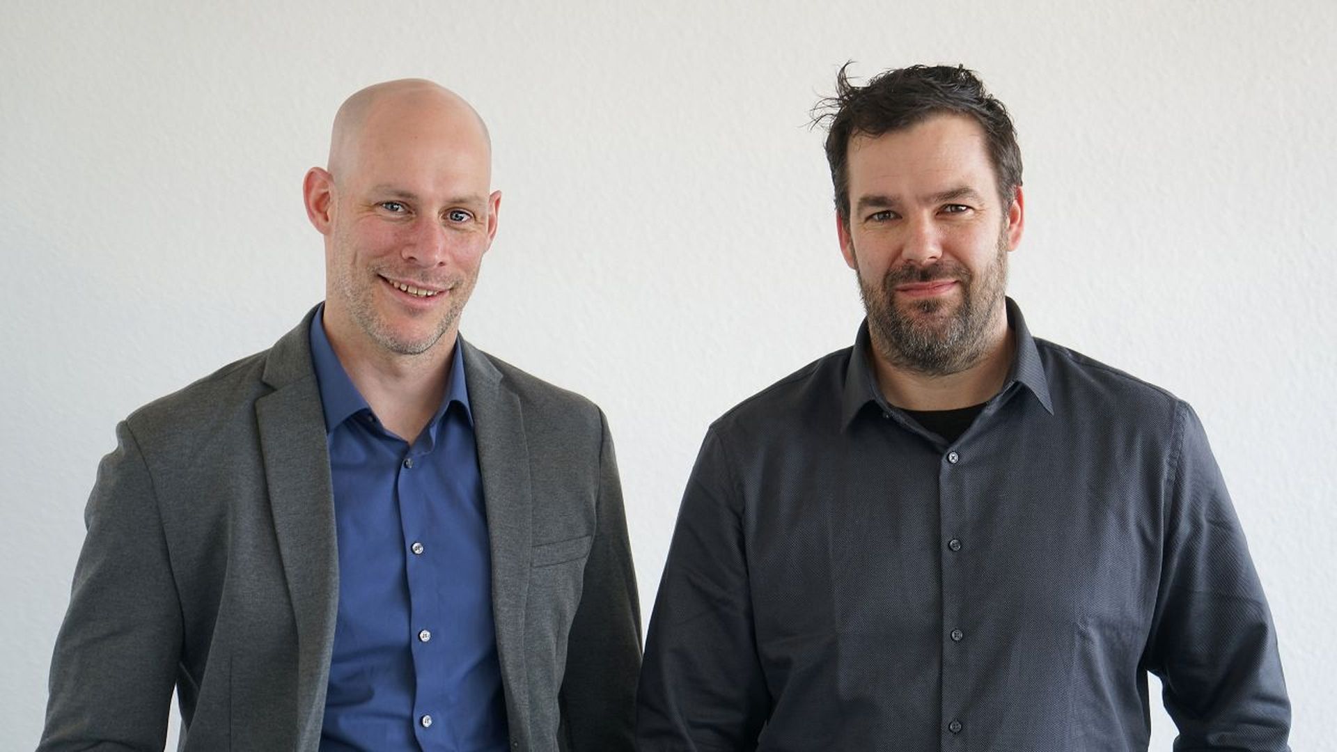 Two men standing side by side, both wearing button-up shirts; one in a gray blazer. They are smiling in front of a plain white background.