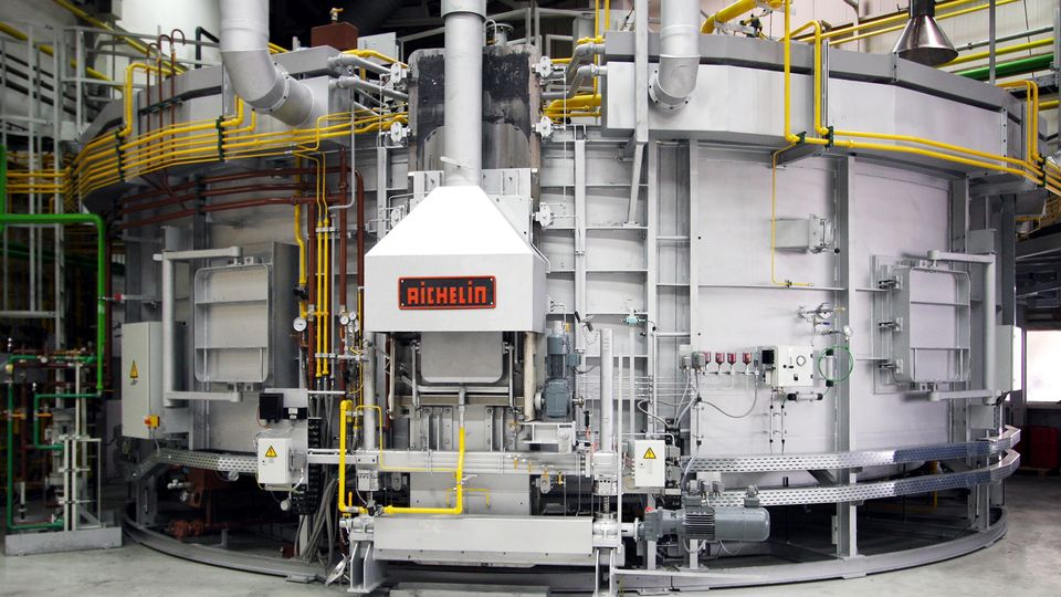 A large industrial furnace with metallic components, yellow pipes, and a central control panel labeled "Aichelin."