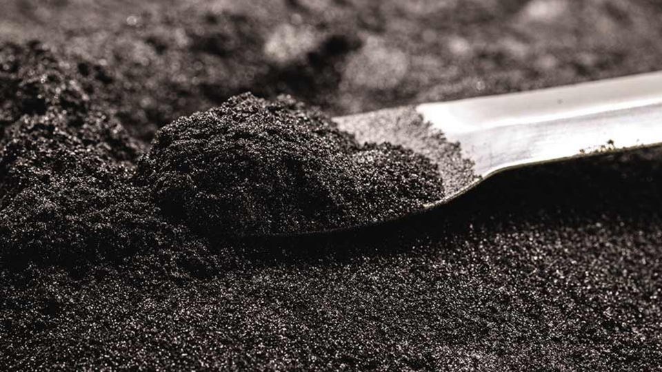 A close-up of a metal spoon scooping fine, black granular powder, resembling activated charcoal, on a textured surface.