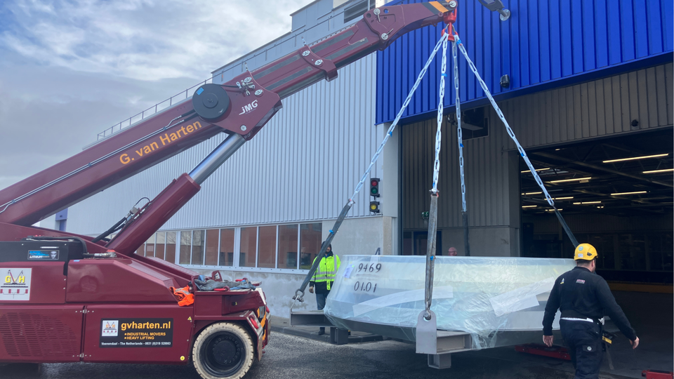 A red industrial crane labeled "G. van Harten" lifts a large, wrapped object outside a warehouse while two workers supervise.