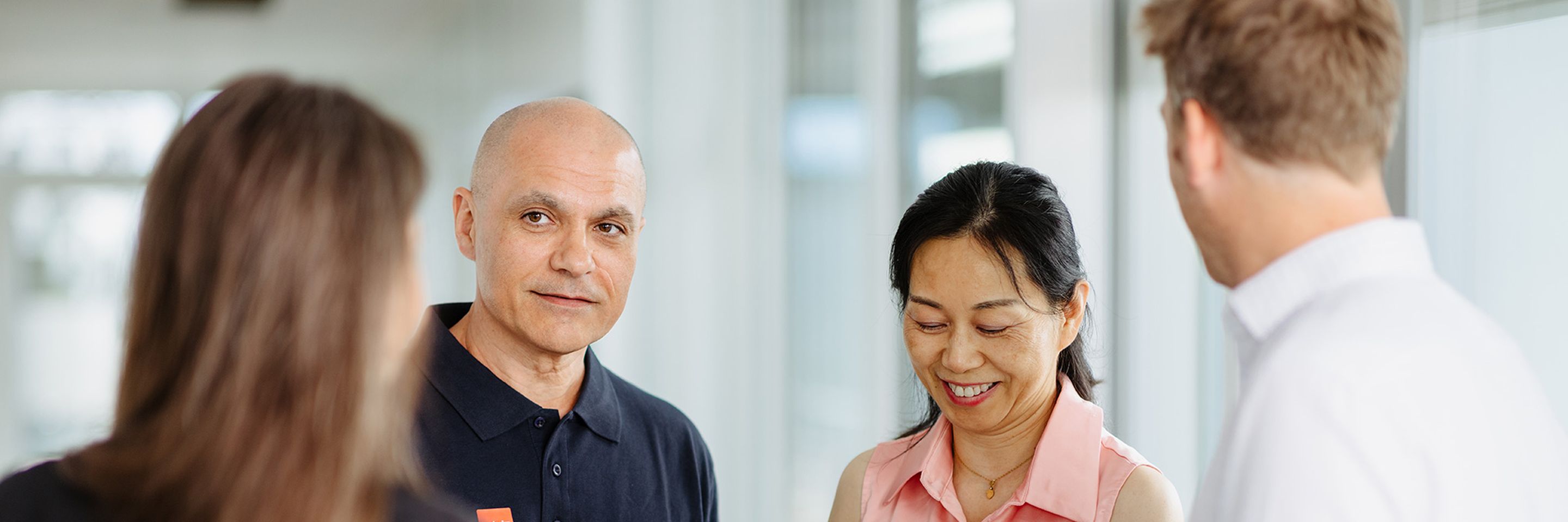 Four people engaged in a friendly conversation, holding a brochure, in a bright office setting.