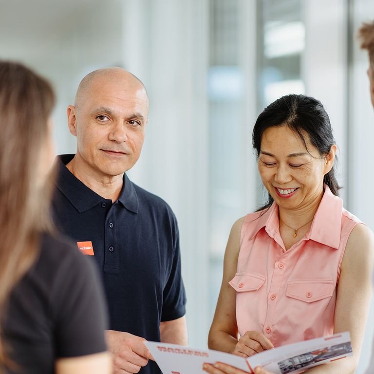 Four people engaged in a friendly conversation, holding a brochure, in a bright office setting.