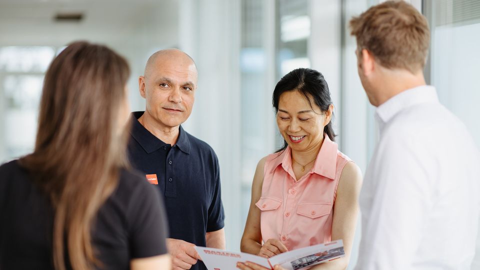 Four people engaged in a friendly conversation, holding a brochure, in a bright office setting.