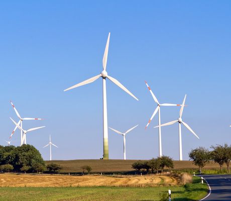 A rural landscape features several large wind turbines on a grassy field under a clear blue sky, with a road curving through the scene.