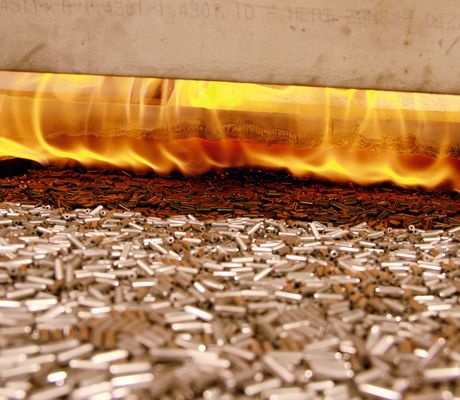 Metal parts on a conveyor belt moving through a furnace with bright flames above, indicating a heating or tempering process.