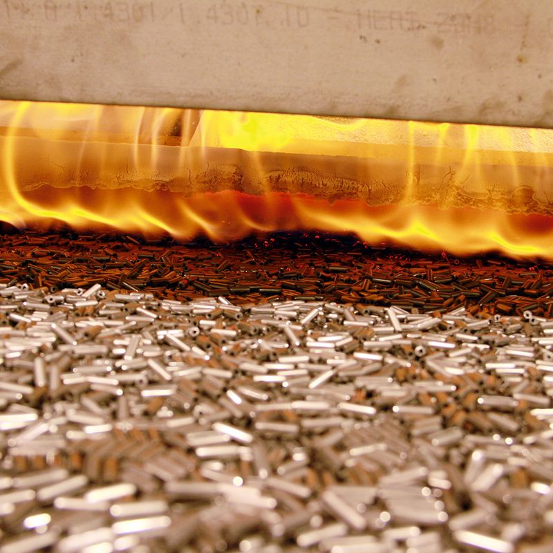 Metal parts on a conveyor belt moving through a furnace with bright flames above, indicating a heating or tempering process.