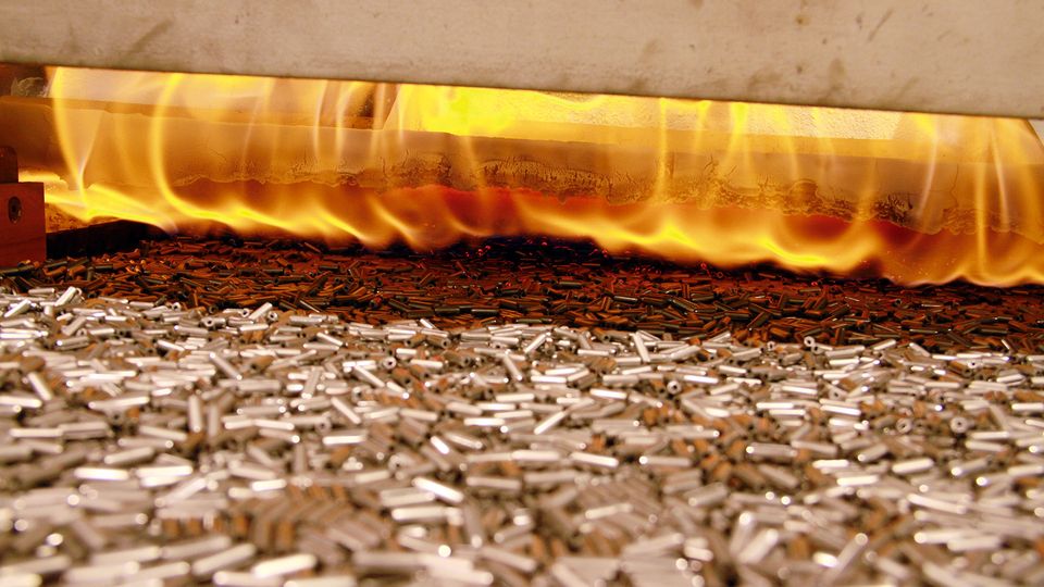 Metal parts on a conveyor belt moving through a furnace with bright flames above, indicating a heating or tempering process.
