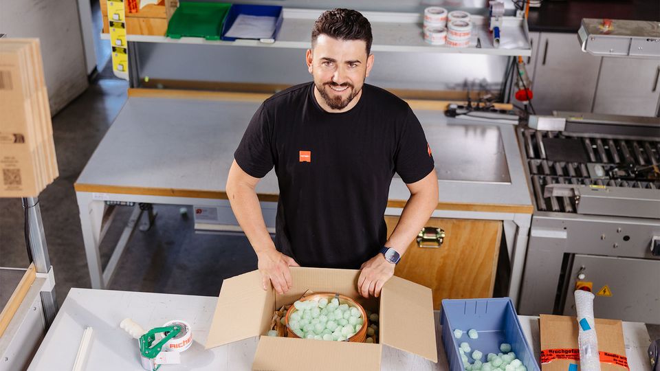 Man packing a cardboard box with protective foam peanuts in a workshop. Various packing materials are on the table.