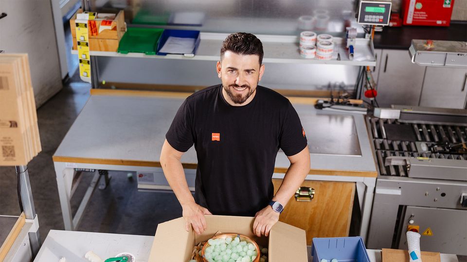 Man packing a cardboard box with protective foam peanuts in a workshop. Various packing materials are on the table.