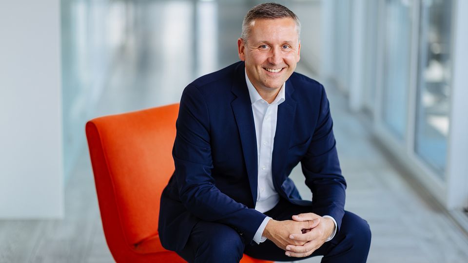 Smiling man in a blue suit sits on an orange chair in a bright, modern office hallway.