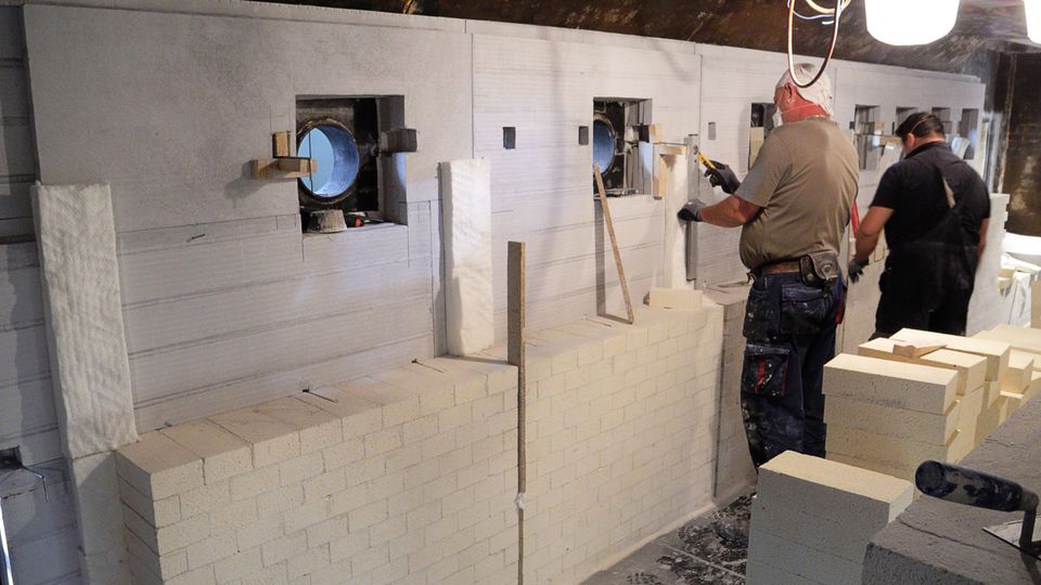 Two workers constructing a wall with light-colored bricks in an industrial setting, with tools and lighting overhead.