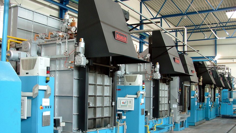 A row of industrial chamber furnaces with blue bases and black hoods lined up in a spacious factory setting with a high, metal ceiling.