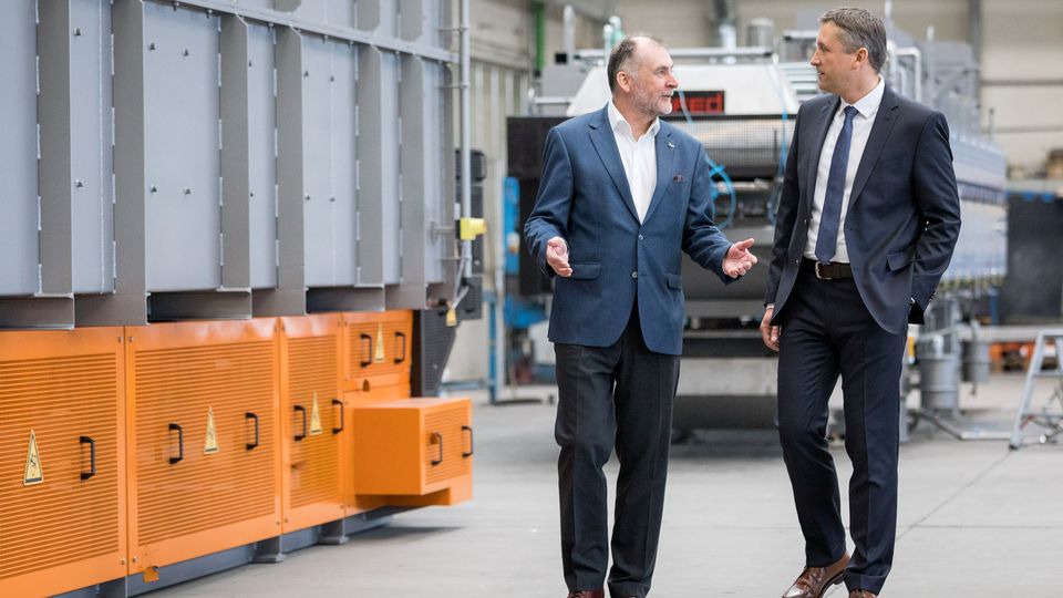 Two men in business attire walk and converse in an industrial setting, surrounded by machinery and metal structures.