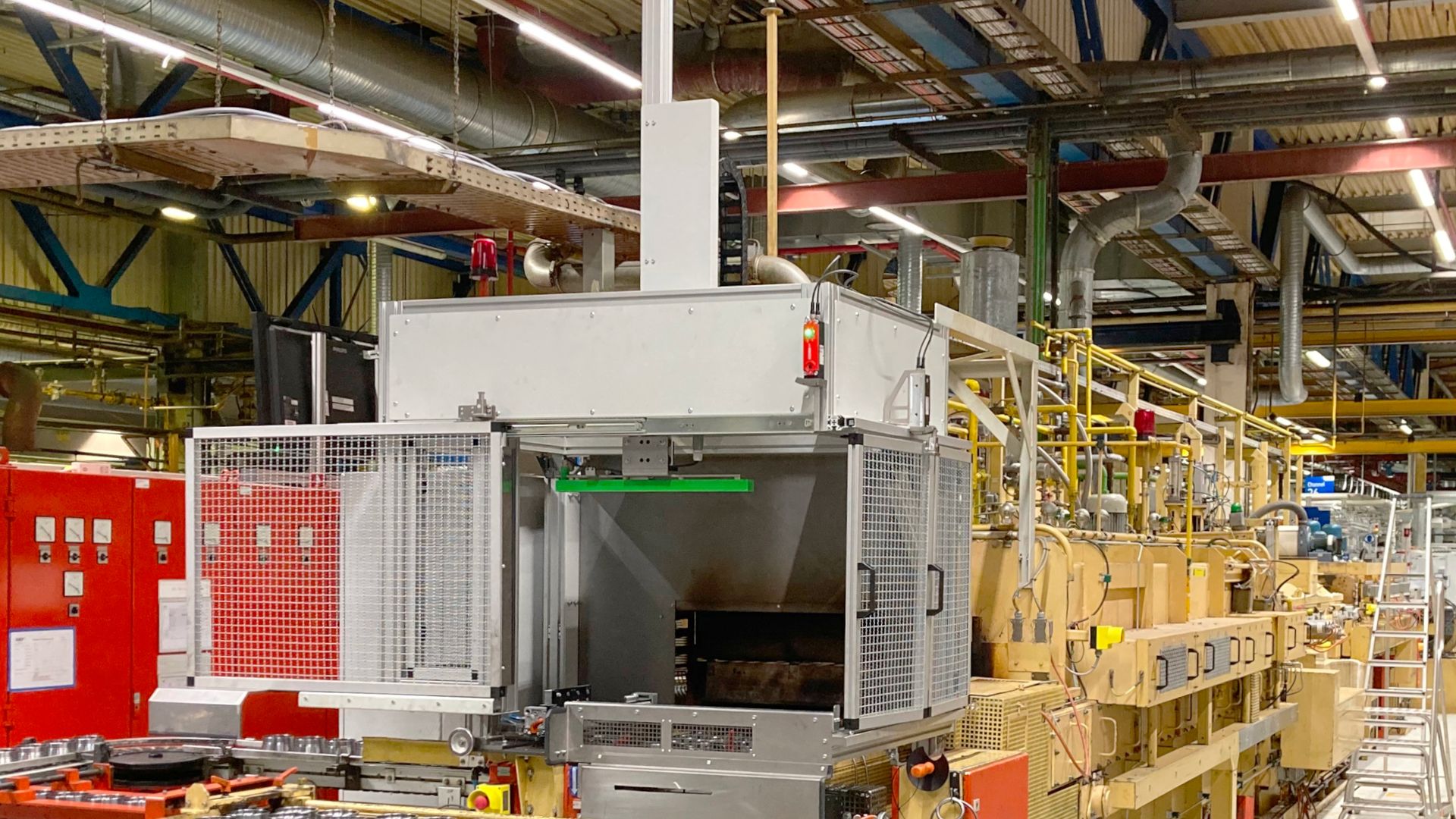 Industrial machinery in a factory setting, with pipes and ceiling rails above. Equipment includes a large metal chamber and control panels.