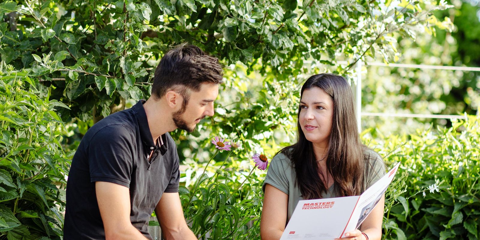 A man and woman sit on a bench, engaged in conversation, as the woman holds a magazine. Lush greenery surrounds them.