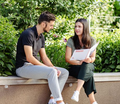 A man and woman sit on a bench, engaged in conversation, as the woman holds a magazine. Lush greenery surrounds them.