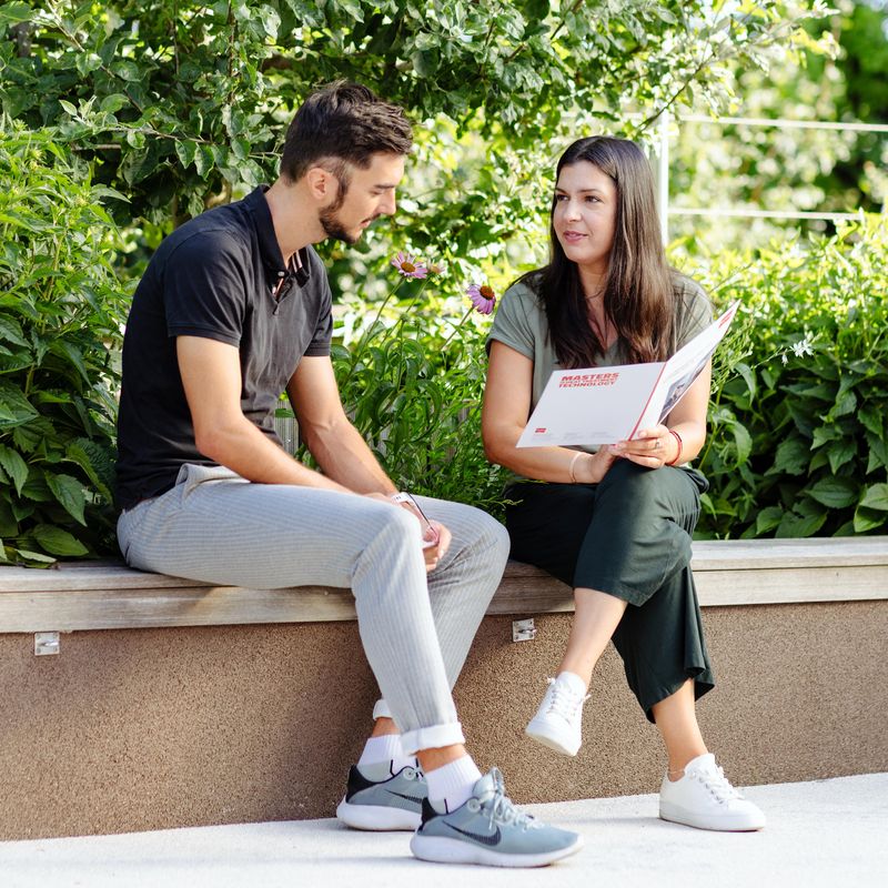 A man and woman sit on a bench, engaged in conversation, as the woman holds a magazine. Lush greenery surrounds them.