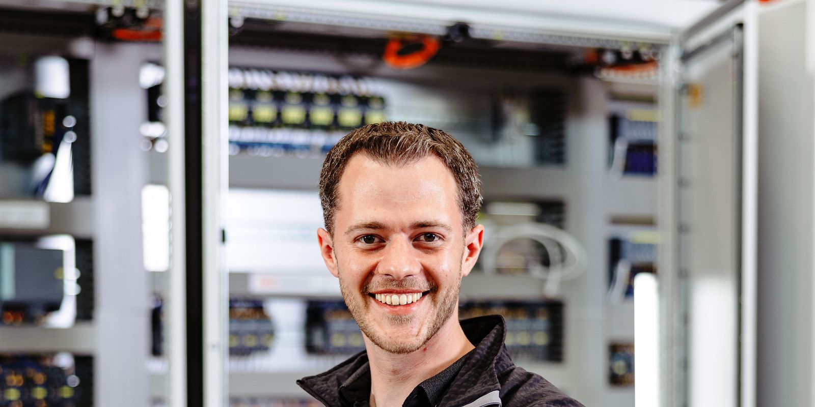 Smiling person in a black jacket stands confidently in front of open electrical panels, with a laptop on a table nearby.