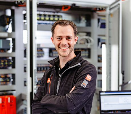 Smiling person in a black jacket stands confidently in front of open electrical panels, with a laptop on a table nearby.