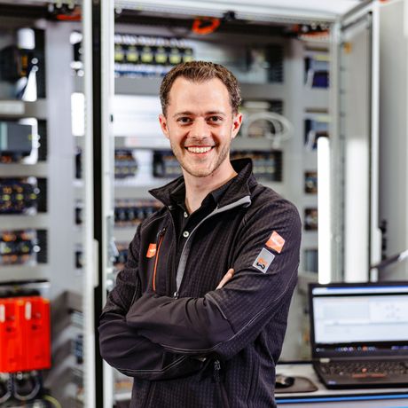 Smiling person in a black jacket stands confidently in front of open electrical panels, with a laptop on a table nearby.