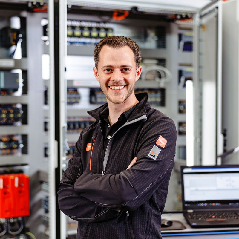 Smiling person in a black jacket stands confidently in front of open electrical panels, with a laptop on a table nearby.