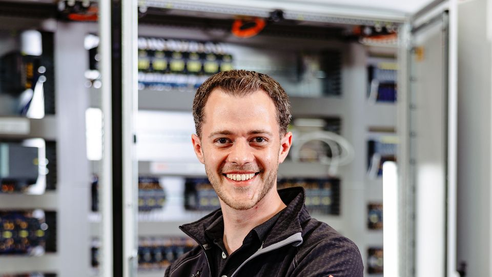 Smiling person in a black jacket stands confidently in front of open electrical panels, with a laptop on a table nearby.