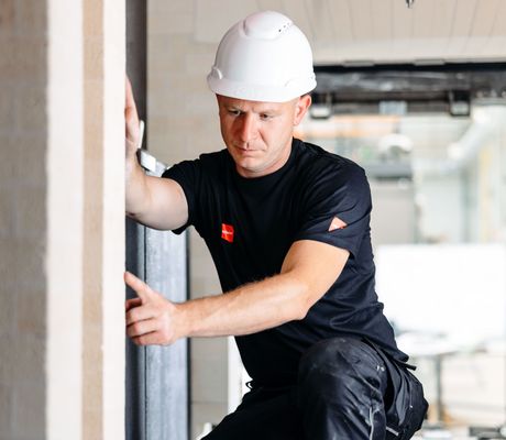 Construction worker in a white hard hat and black outfit assessing a wall indoors. Building materials are visible in the background.