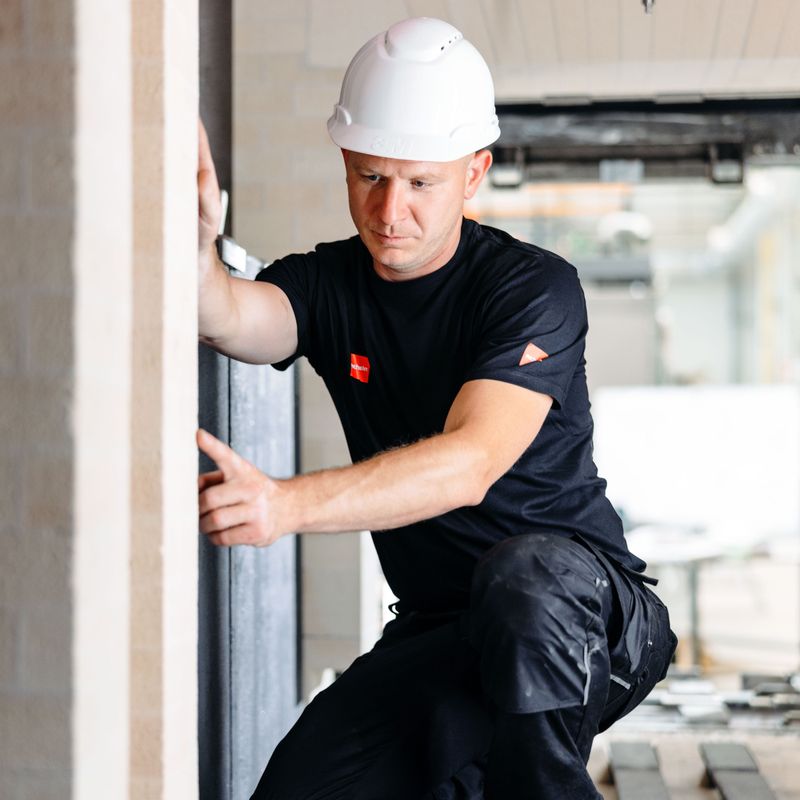 Construction worker in a white hard hat and black outfit assessing a wall indoors. Building materials are visible in the background.