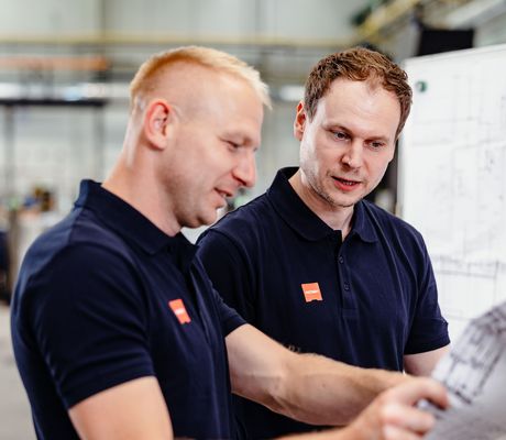 Two men in navy shirts collaborate, examining blueprints in an industrial setting. They appear focused and engaged in their discussion.