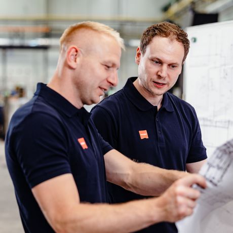 Two men in navy shirts collaborate, examining blueprints in an industrial setting. They appear focused and engaged in their discussion.