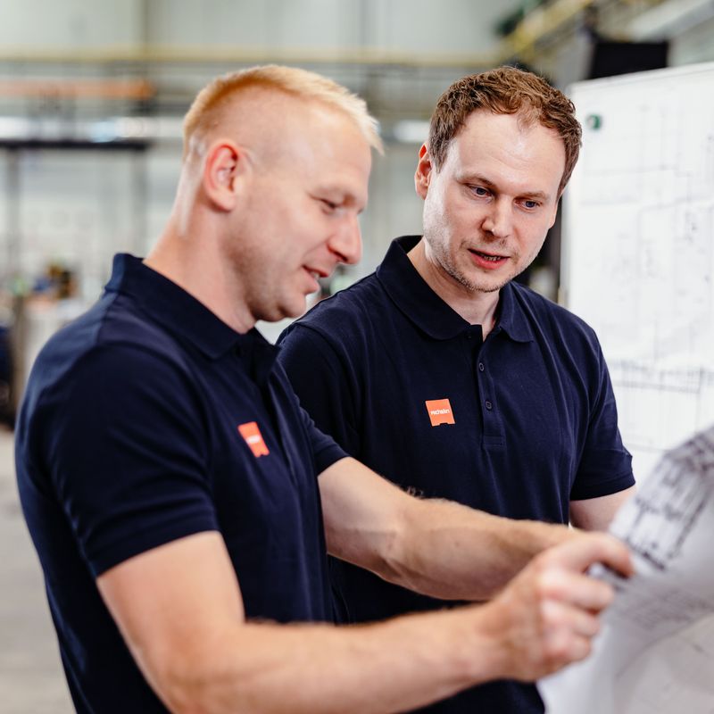 Two men in navy shirts collaborate, examining blueprints in an industrial setting. They appear focused and engaged in their discussion.