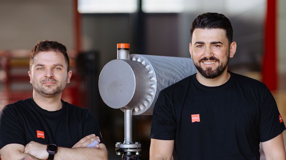 Two service men in black shirts posing next to industrial equipment, one standing with crossed arms and the other sitting, in a workshop setting.