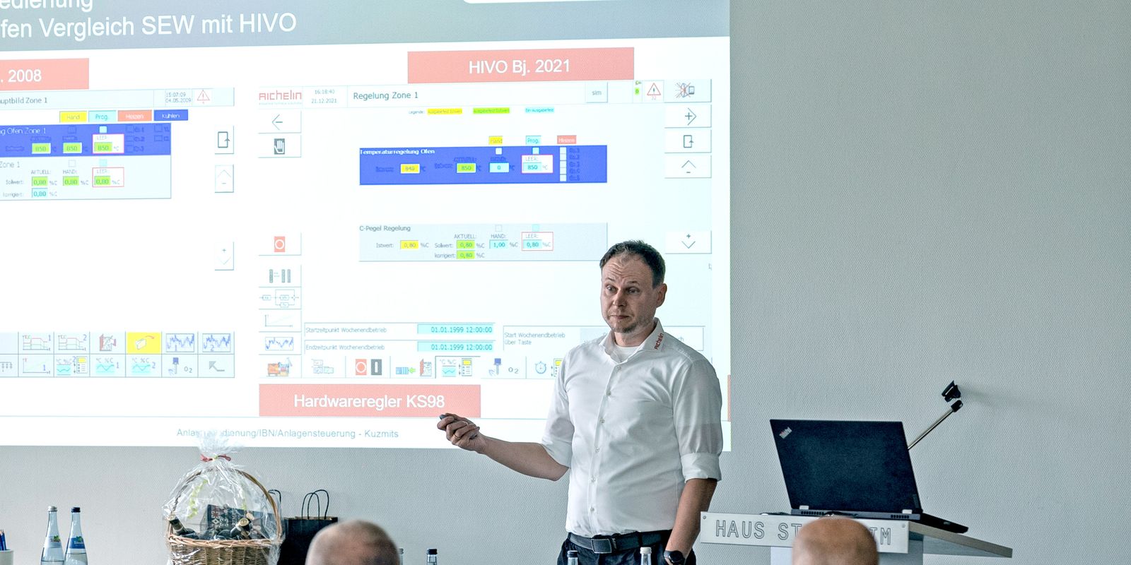 A person giving a presentation in a conference room, with a projected slide showing graphs and text. Attendees are seated and attentive.