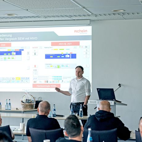 A person giving a presentation in a conference room, with a projected slide showing graphs and text. Attendees are seated and attentive.
