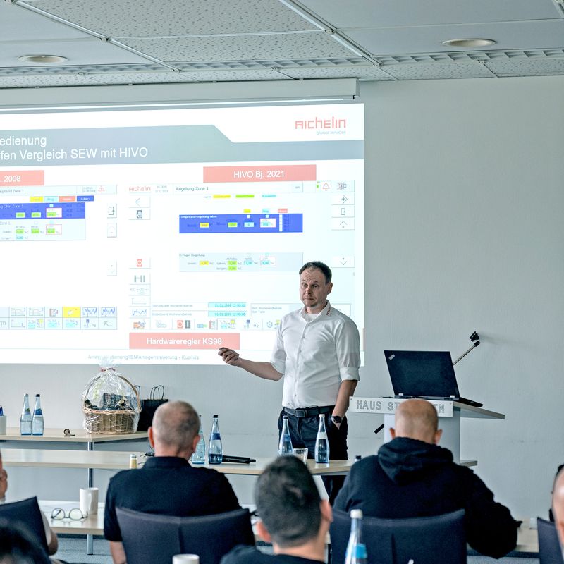 A person giving a presentation in a conference room, with a projected slide showing graphs and text. Attendees are seated and attentive.