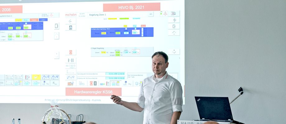 A person giving a presentation in a conference room, with a projected slide showing graphs and text. Attendees are seated and attentive.