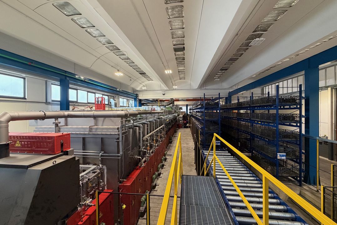 Interior of an industrial facility with metal machinery, pipes, a walkway with yellow railing, and metal shelving in a large, well-lit space.