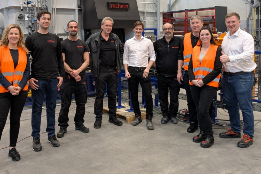 A group of people in a workshop, some wearing orange safety vests, stand together smiling in front of machinery. DAF Trucks and Aichelin