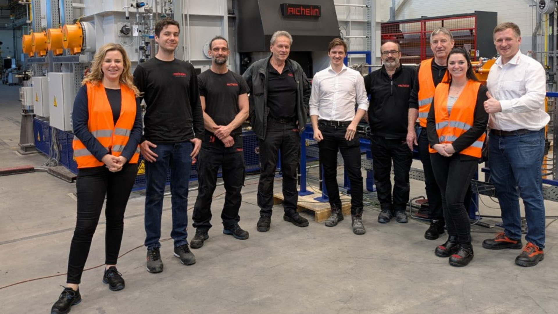 A group of people in a workshop, some wearing orange safety vests, stand together smiling in front of machinery. DAF Trucks and Aichelin