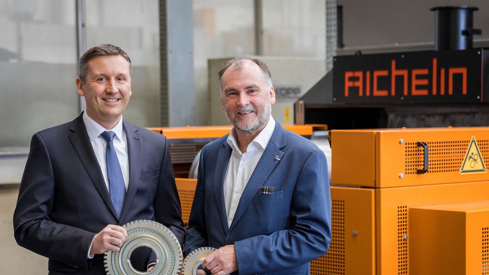 Two men in suits holding large metal gears, standing in front of industrial orange machinery marked "AICHELIN," indoors.