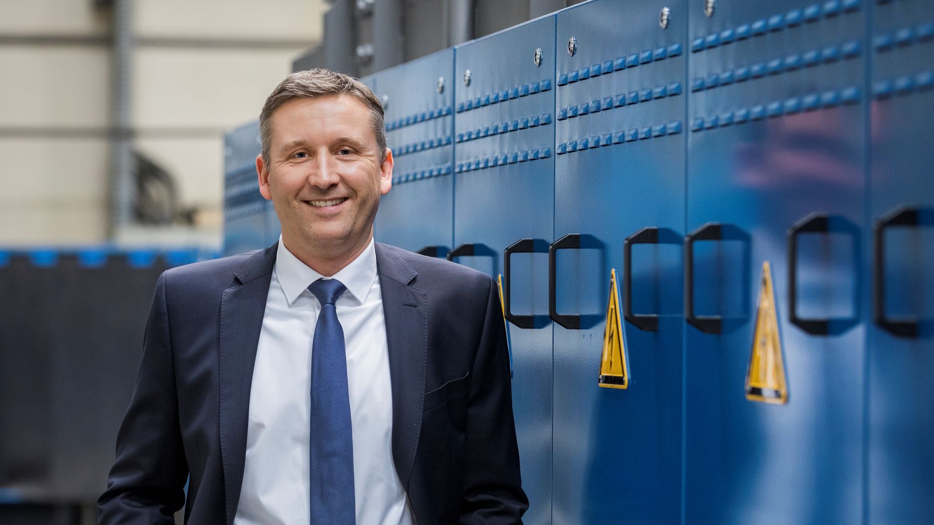 A man in a suit stands smiling next to industrial blue machinery, hands in pockets, in a professional setting.