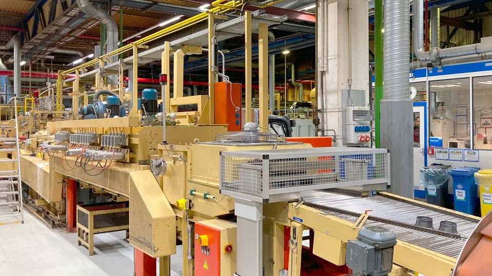Industrial machinery in a factory setting, featuring conveyor belts, control panels, and overhead pipes, under a high ceiling.